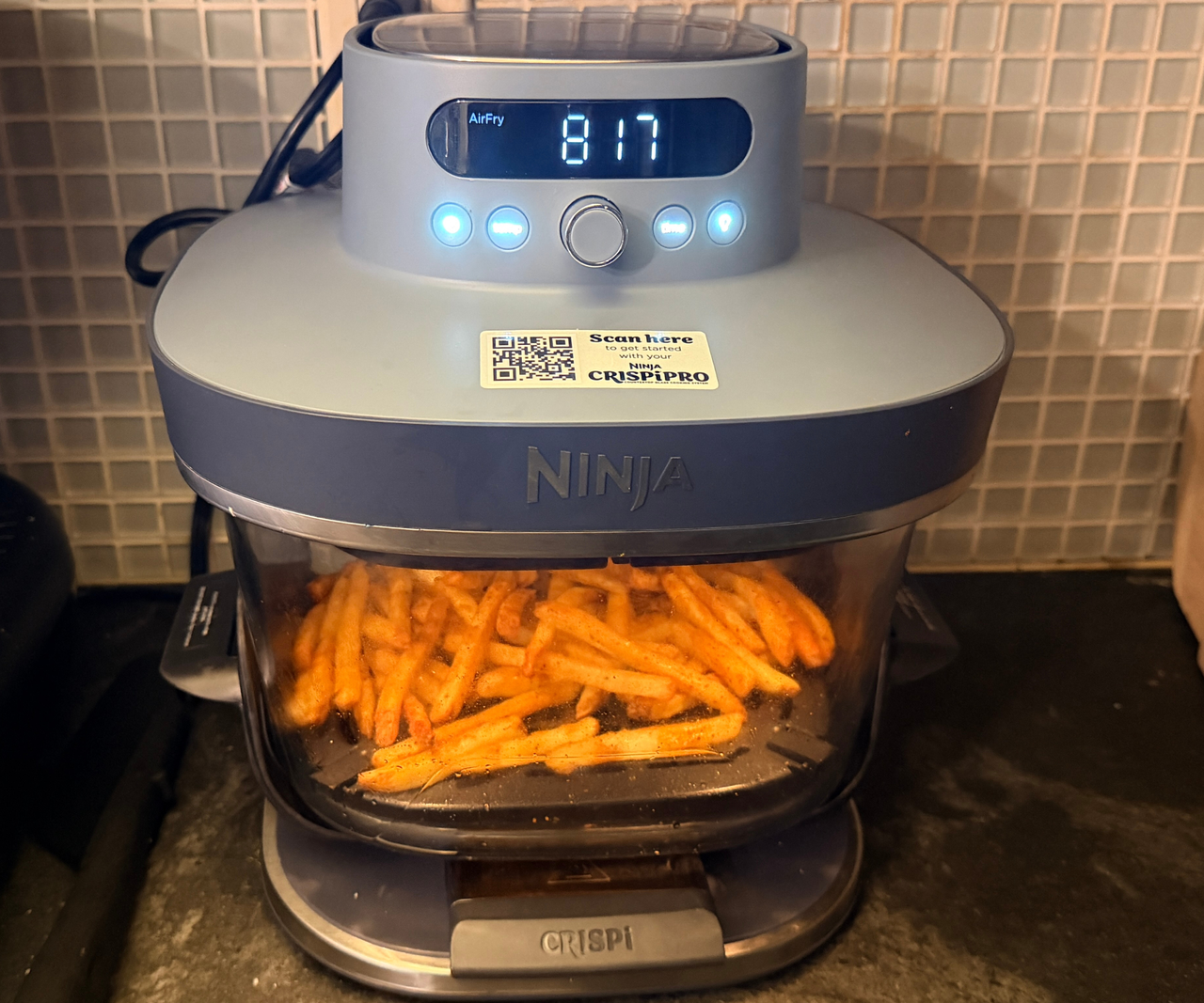 A glass Ninja Crispi Pro air fryer base under the blue air fryer head on a black kitchen counter. The glass dish is filled with fries.