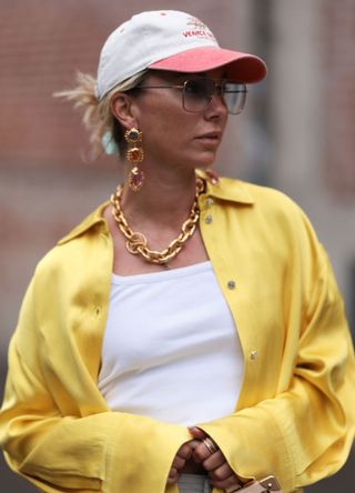 Karin Teigl seen wearing Andy Wolf light blue transparent pilot sunglasses, red / white baseball cap, gold earrings and statement chain necklace, white cotton top, Zara grey striped nylon wide leg pants, Stine Goya yellow silk blouse shirt, Louis Vuitton brown leather / logo pattern canvas handbag, during the Copenhagen Fashion Week Spring/Summer 2024 on August 09, 2023 in Copenhagen