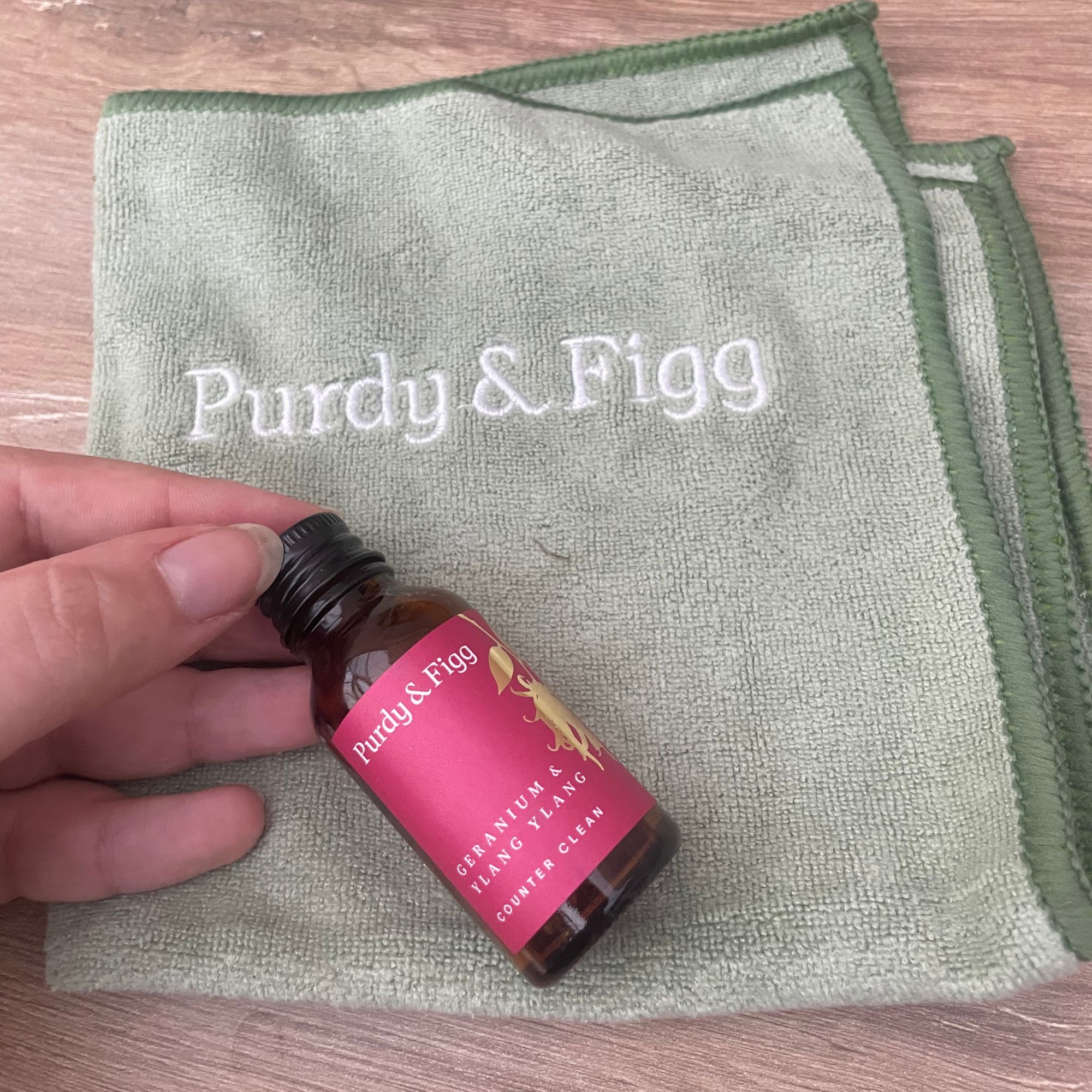 Hand holding Purdy &amp;amp; Figg refill bottle on top of a green, branded cleaning cloth