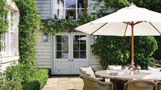White outdoor umbrella shading a round outdoor dining table and chairs, which sit on a charming stone patio with crawling vines