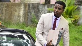 Sterling Fox (Trieve Blackwood-Cambridge) in Death in Paradise season 14 episode 7. He is wearing a cream-coloured suit with a white shirt and a red tie. He is getting out of his open-topped sports car and carrying several case files in his right hand.