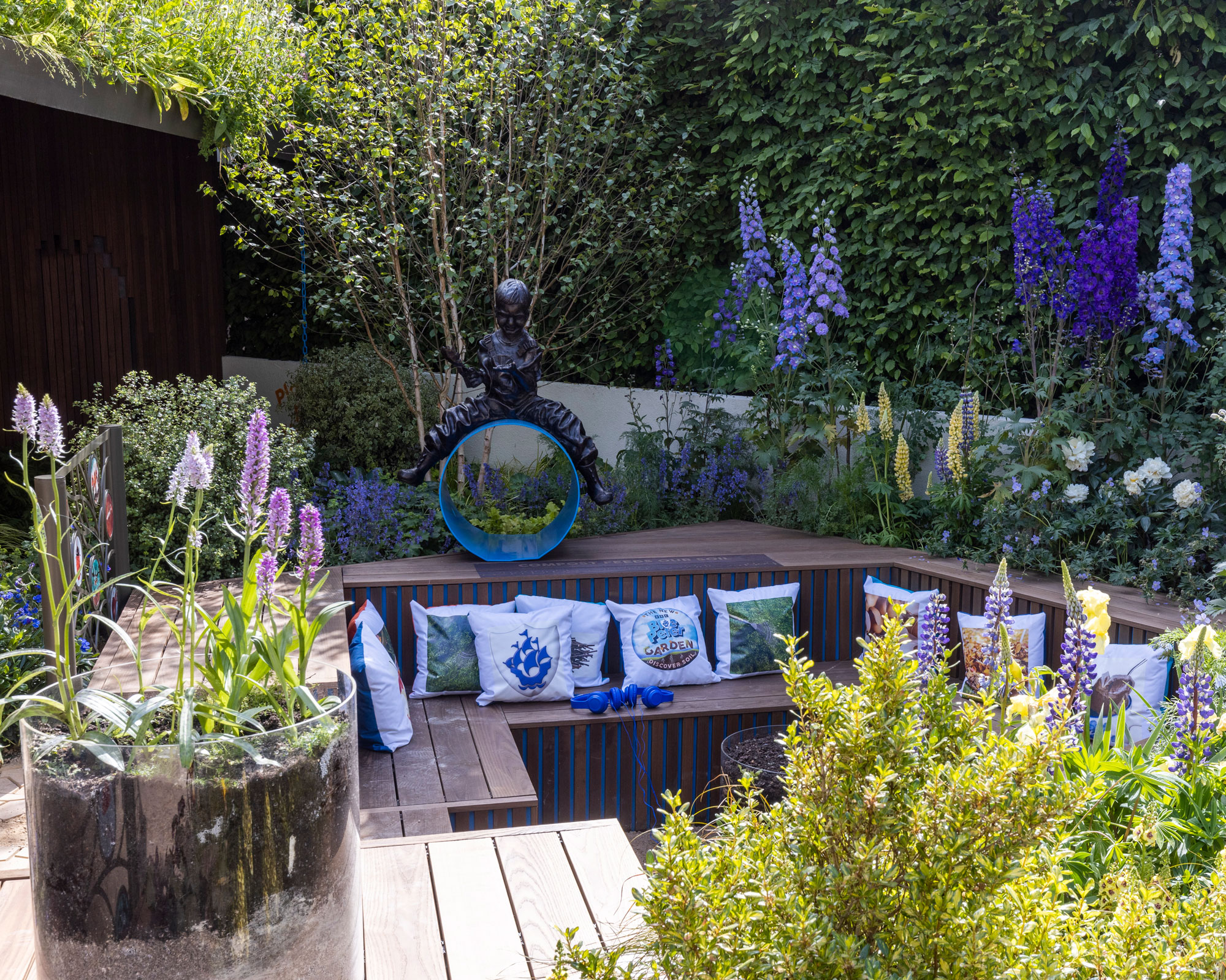 sunken conversation pit design with benches and cushions, surrounding by delphiniums and lupins, with a mirror, statue and glass container with more flowers