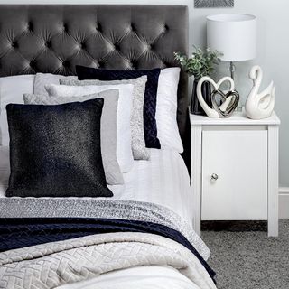 Grey and white bedroom with bed linen in different shades of grey, a white side table with white and silver ornaments and a lamp