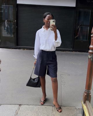 Swedish influencer Sylvie Mus wearing a white button-down shirt, tailored Bermuda shorts, Manolo Blahnik kitten-heel mules, and a black shoulder bag in Paris