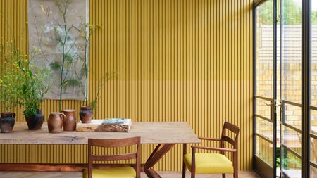 Dining room with large mid century dining table and chairs with yellow seat cushions, yellow paneled walls, large art work canvas, plants on table and large glass doors on right