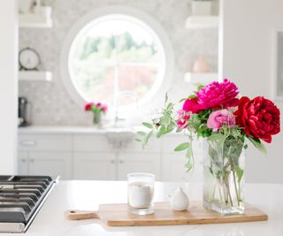 A small white candle on a board next to a vase of pink flowers in a white kitchen