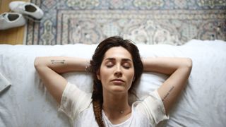 Bedtime stories for adults: A woman with dark hair and wearing a cream t-shirt relaxes on her bed while listening to a bedtime story before sleep