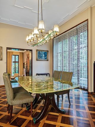 View of the dining room with a Brazillian onyx table and set of patterned chairs by Osvaldo Borsani. There is wooden flooring, tall windows with curtains, a pendant light with ten lights, wall art and a partial view of another room through a rectangle arch