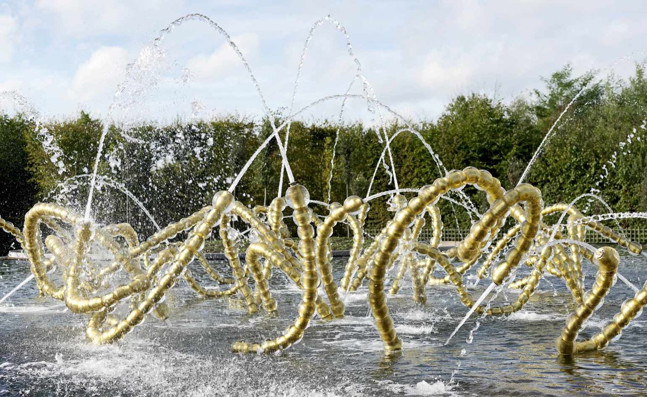 Daytime, outside image, gold and glass decorative piping garden water feature, trees in the backdrop, blue cloudy sky