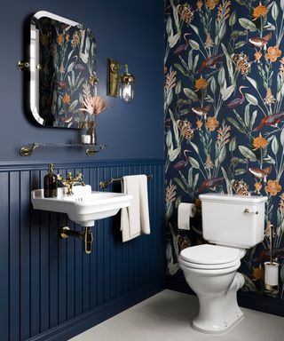 Elegant small bathroom with bold navy wainscoting, tropical-patterned wallpaper, and brass accents. Featuring a compact wall-mounted sink, traditional white toilet, and gold fixtures