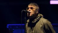 Liam Gallagher wearing sunglass and a green raincoat while singing onstage in Oasis: Don't Look Back In Anger.