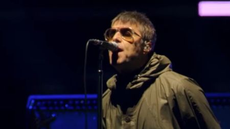 Liam Gallagher wearing sunglass and a green raincoat while singing onstage in Oasis: Don't Look Back In Anger.