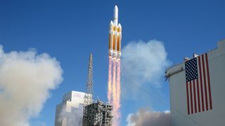 a large white-and-orange rocket lifts off behind an american flag