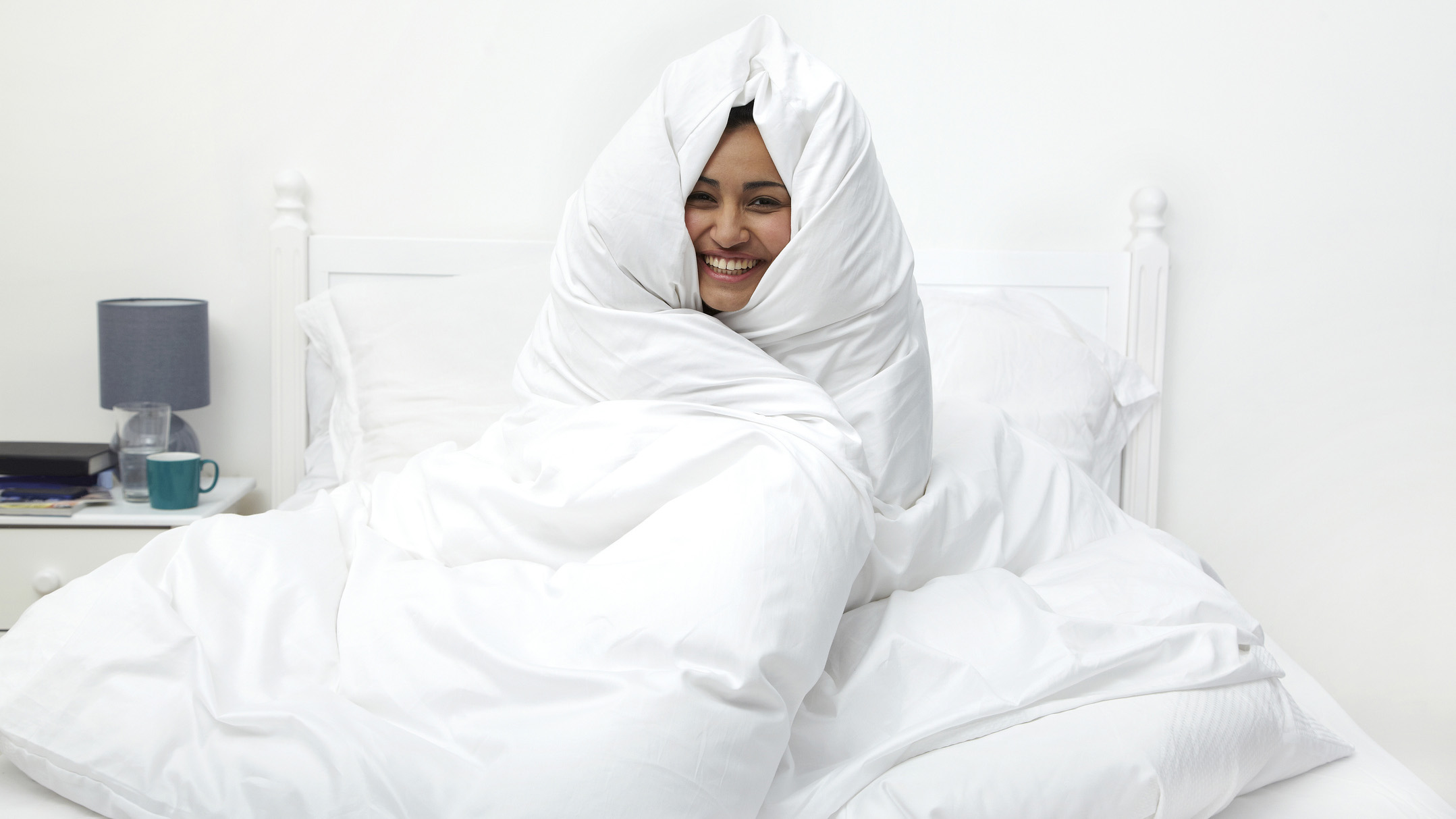 A woman wraps herself from head to toe in a white duvet while laughing, sat on the bed