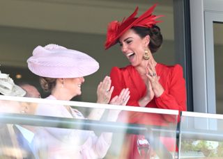 Princess kate standing behind a clear barrier wearing a red hat and dress cheering and clapping while smiling at Duchess Sophie, who is wearing a pink hat and matching dress