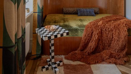 A modern wooden bedframe and headboard with an avocado green sheet, a brown fluffy throw draped over the end and a checkerboard side table sitting in front by the window with patterned curtains