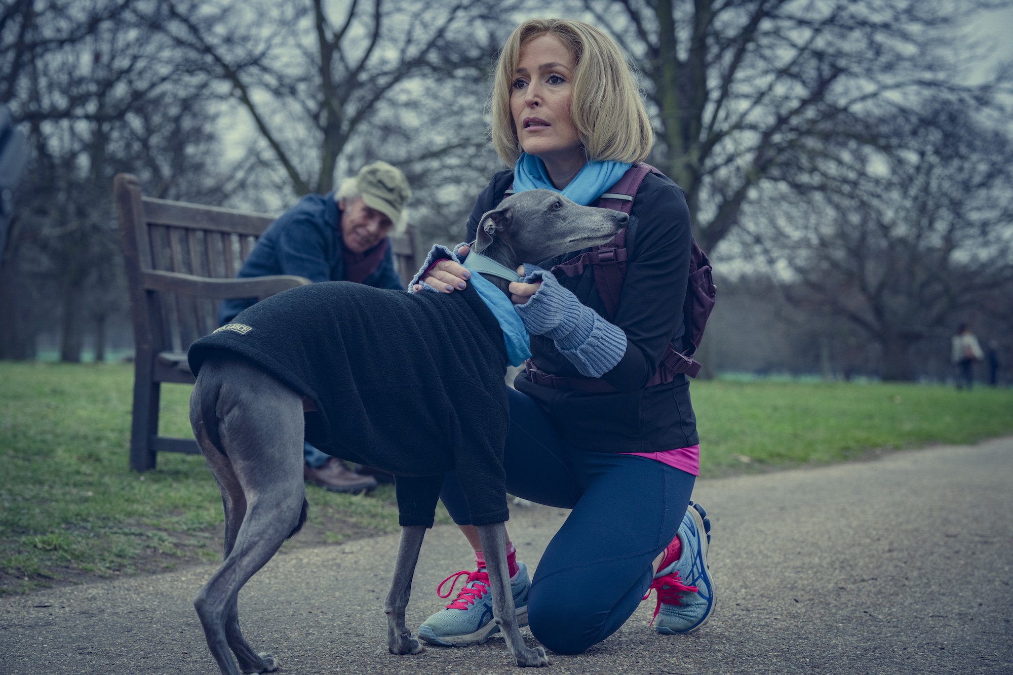 a woman in running gear (gillian anderson as emily maitlis kneels and holds her dog's collar, while a man sits on a bench behind her, in the netflix movie 'scoop'