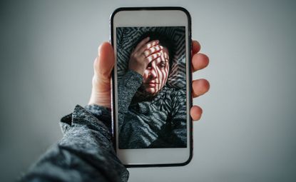 Cropped Hands Of Woman Holding Mobile Phone While Taking Selfie Against Grey Background. Is Instagram bad for mental health?