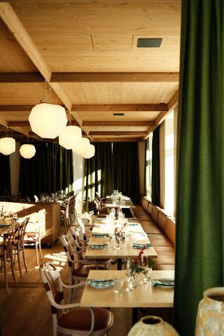 A sun-kissed hotel restaurant featured wooden interiors punctuated by geometrical pendant lights in yellow, leather banquettes, green curtains, and organic sculptures.