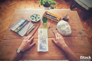 A wooden desk with watercolour paints, brushes, a mask and an artist's hands