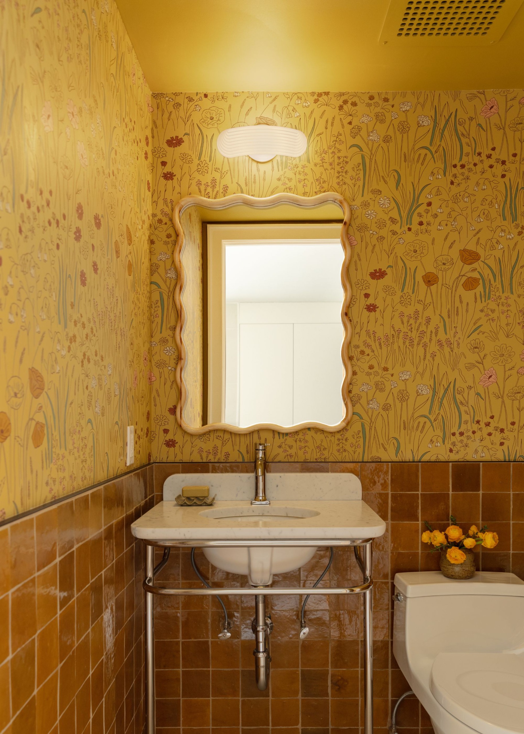 A small powder room with square ochre wall tiles on the lower wall and a sunny yellow whimsical wallpaper on the upper wall. A yellow ceiling, a wavy mirror, and a white sink with chrome hardware.