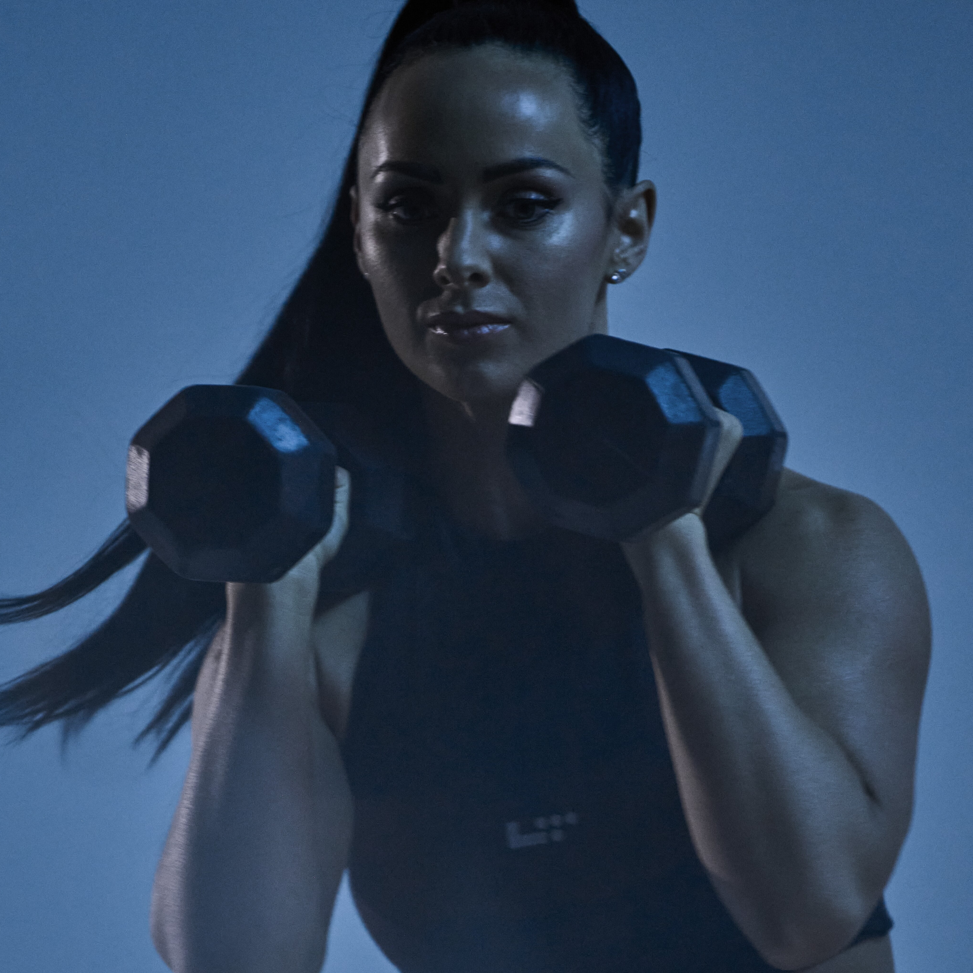 Woman with long black hair in pony tail holds hex dumbbells by her shoulders. She is against a blue background and lit with a blue light.