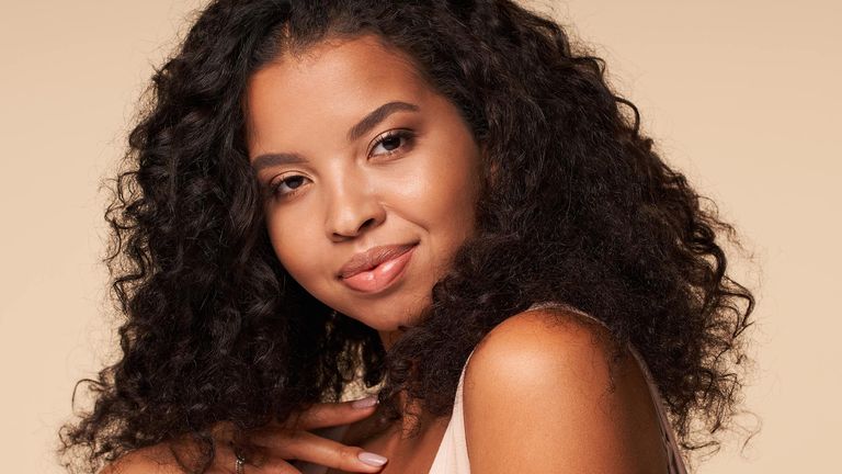 A young woman with curly hair is pictured, she had bright glowing skin and is smiling and seemingly carefree