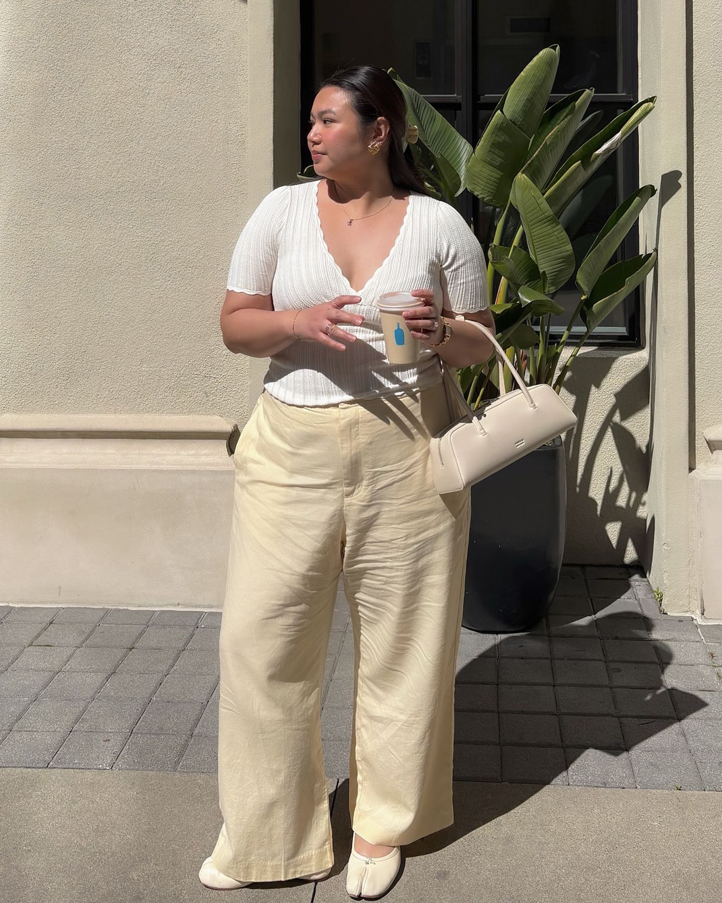 fashion influencer Marina Torres poses on a sidewalk next to a palm pant wearing a white ribbed v-neck top, ivory east-west bag, butter-yellow Gap linen pants, and ivory white Maison Margiela flats