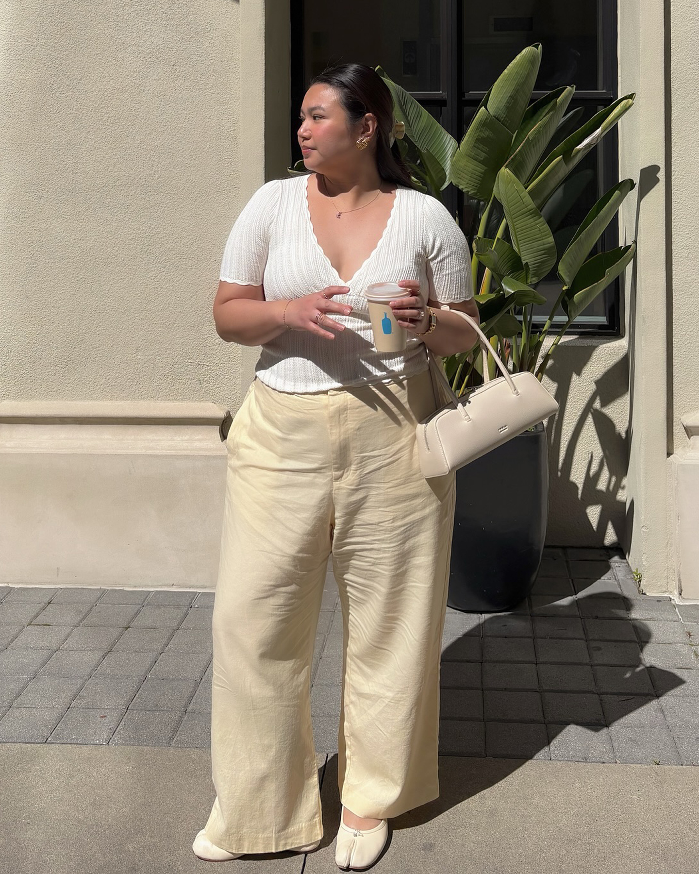 fashion influencer Marina Torres poses on a sidewalk next to a palm pant wearing a white ribbed v-neck top, ivory east-west bag, butter-yellow Gap linen pants, and ivory white Maison Margiela flats