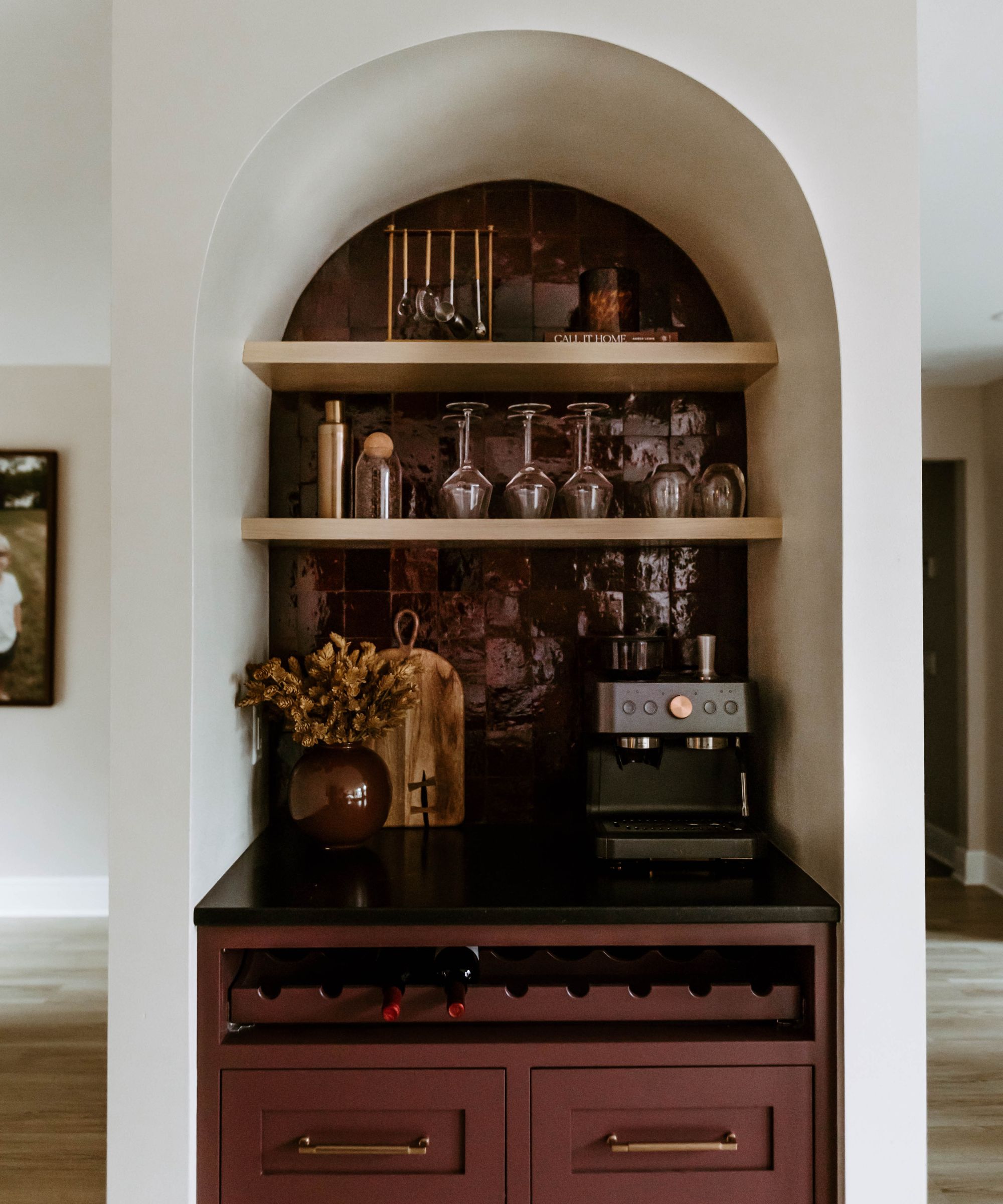 A small home bar installed into an arched alcove with wine red glossy tiles and cabinetry paired with brass shelving