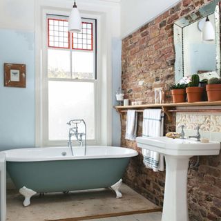 bathroom with blue freestanding bath, exposed brick wall, white basin and wooden shelving with plant pots