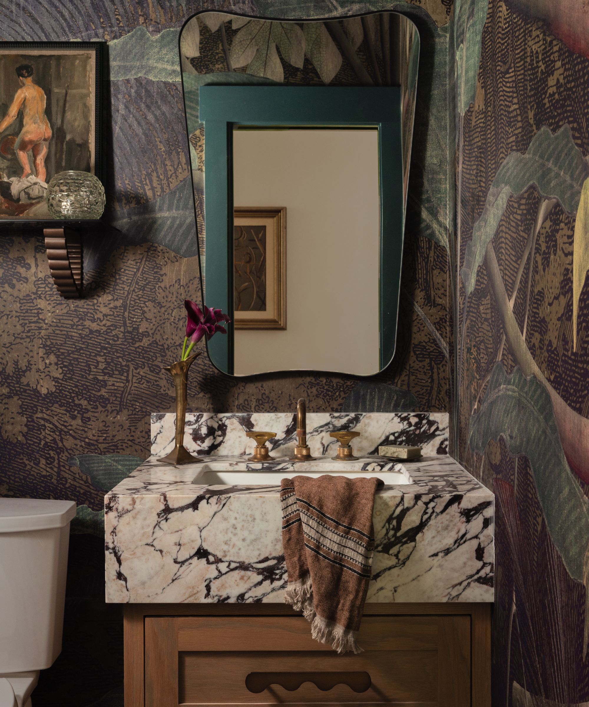 a botanical wallpaper in a powder room with a small wooden vanity, large veined marble sink and a curved mirror