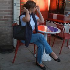 Kathryn Zahorak sitting at a table with a sweater over her shoulders wearing a woven bag, white t-shirt, jeans, and black ballet flats