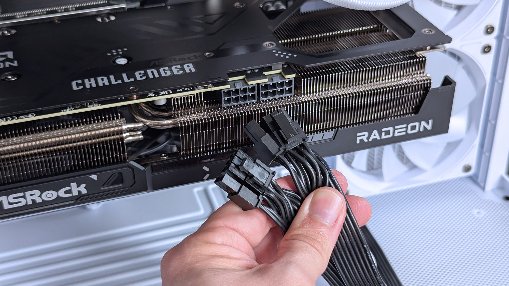 A hand holds black power cables near an ASRock Challenger Radeon RX 9070 XT GPU inside an iBUYPOWER RDY Trace X R01 pre-built gaming PC with a white case