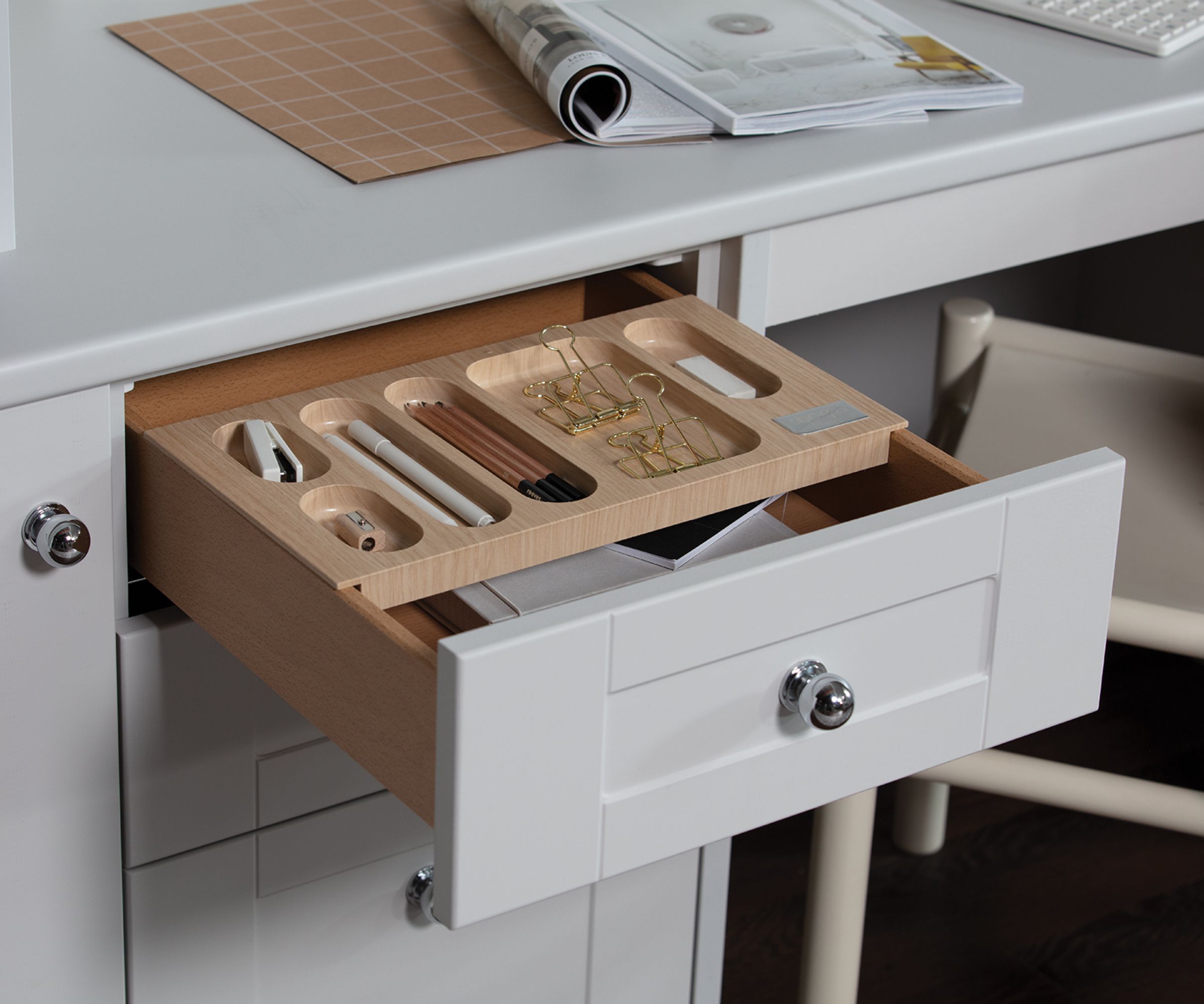Open white desk drawer with a wooden organiser tray neatly holding pens, pencils, clips, and a stapler; magazine and keyboard visible on the desk above.