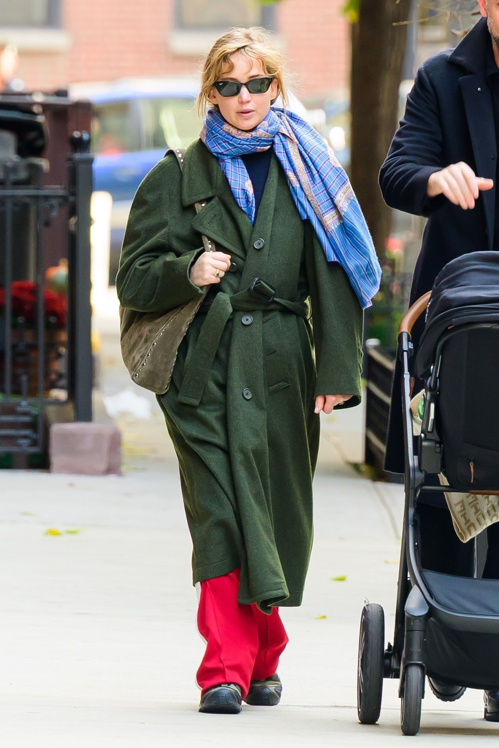 Jennifer Lawrence walks down the street in New York wearing red trousers with an olive green coat and blue scarf.