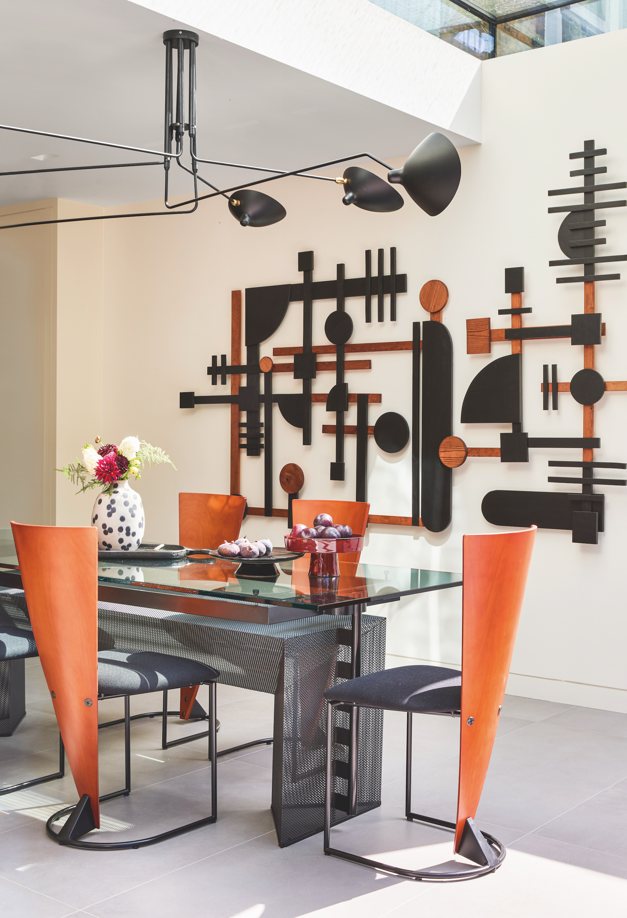 Dining are with an abstract, geometric art piece on the wall behind. The dining table is glass and the dining chairs are a combination of black metal and orange toned wood.
