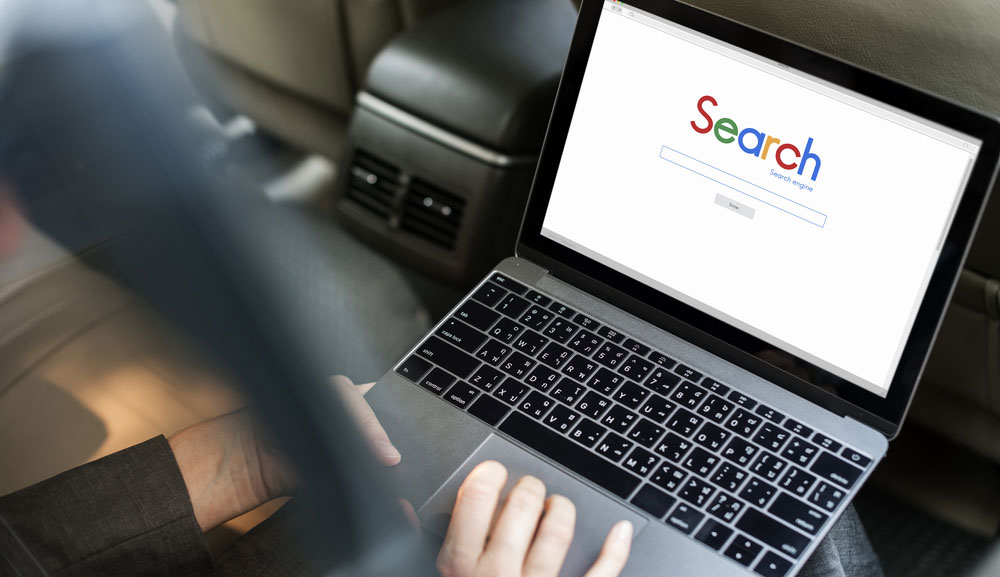 Person using a laptop in the back seat of a car with generic search engine on laptop screen.