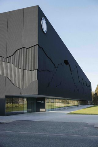 Image of Mountblanc Haus, lawn to front of building, mirrored wall leading to black door entrance, grey stone paved pathway, blue sky