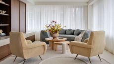 modern living room with gray curved sofa in front of two windows covered in sheer curtains. a timber nesting coffee table is styled with a vase of flowers, and two boucle armchairs sit on the other side. built-in cabinetry in walnut is a mix of open and closed shelves