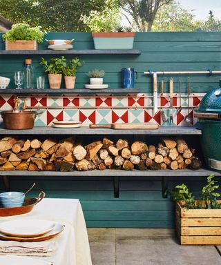 Outdoor kitchen cost on a budget, illustrated by a blue painted fence with wall mounted shelving housing logs, a sink, kitchen crockery and colorful tiles.