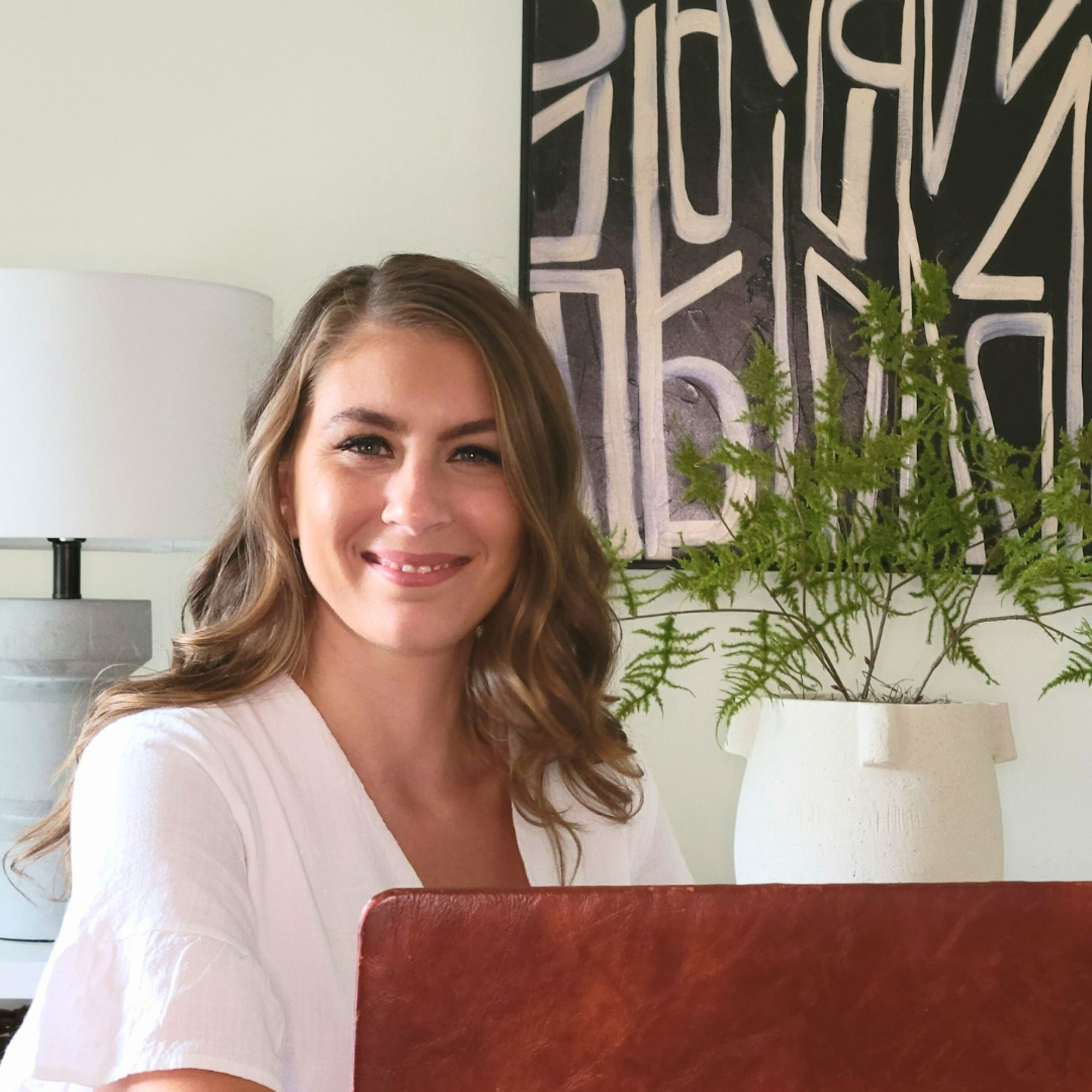 A picture of Kristin Patrician, a brunette woman in a white t-shirt sitting in front of a red brown leather laptop, with green foliage , black and white artwork, and a gray and white ceramic lamp behind her