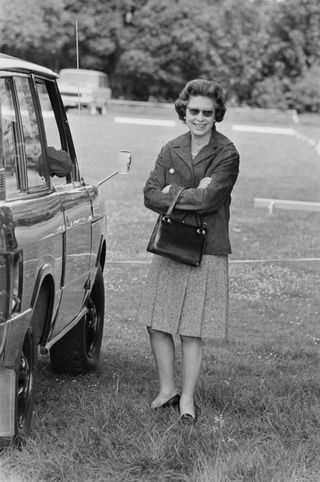 A black and white photo of Queen Elizabeth wearing a skirt and standing next to her Land Rover outside