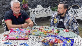 Paul Hollywood in a black t-shirt sits at a white table covered in vibrant sweets, alongside piñata creator Dalton who is wearing a camouflage jacket in Paul Hollywood Eats Mexico.