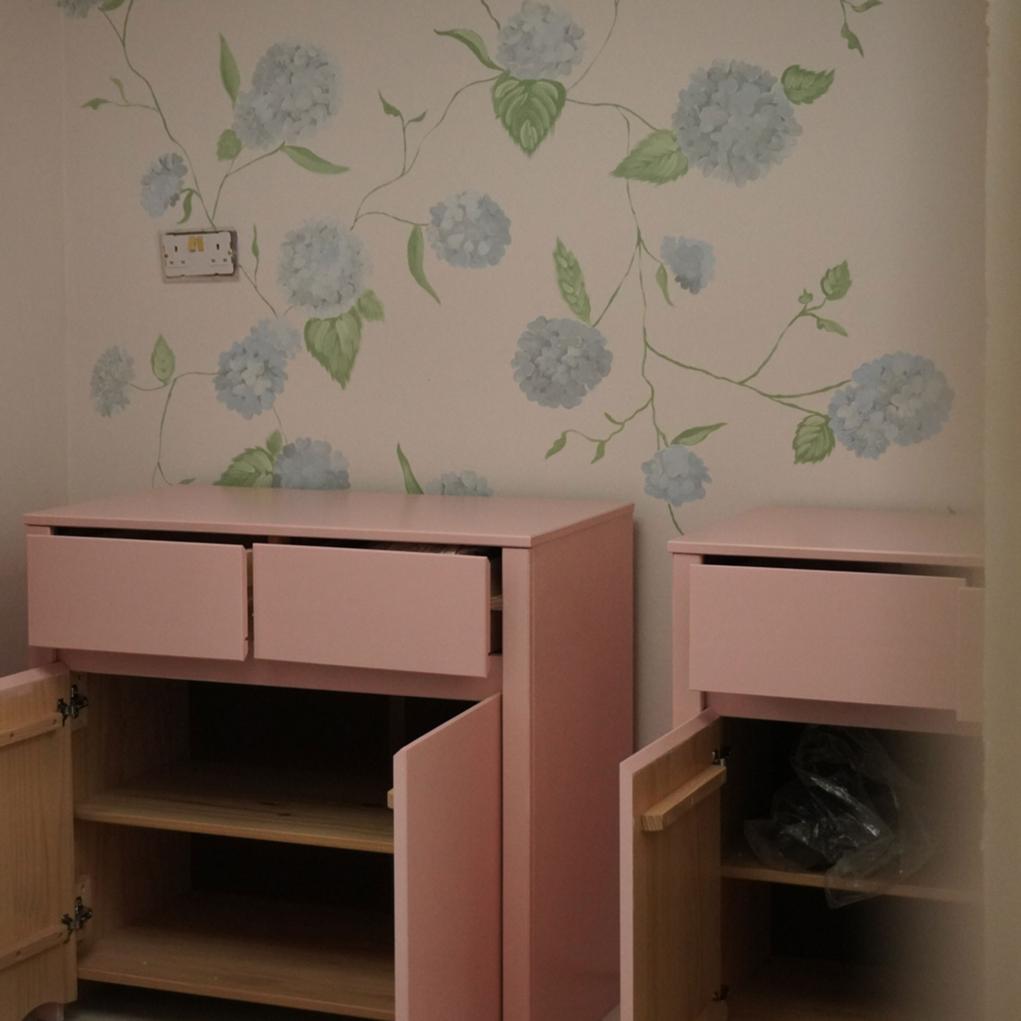Two pink cabinets with open doors sitting in front of wall painted with blue hydrangea pattern