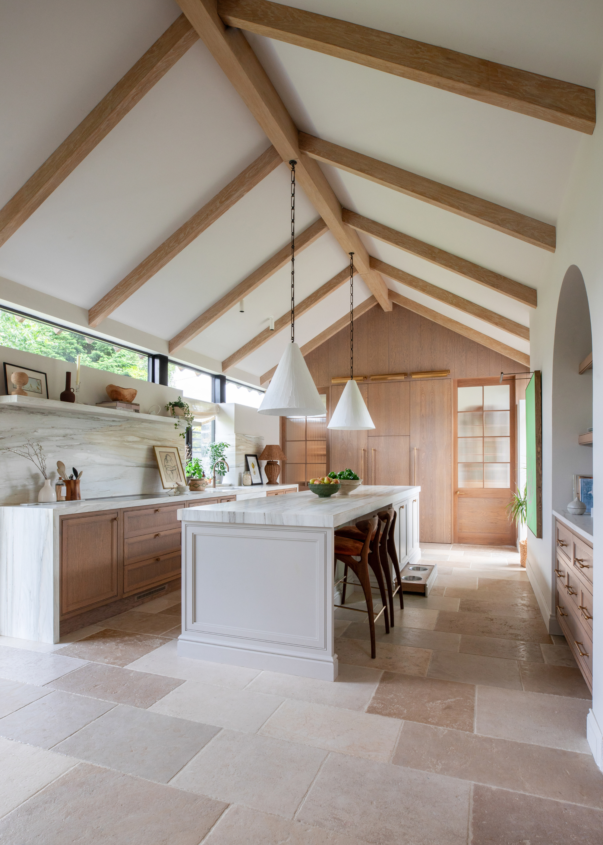 A large kitchen with a vaulted ceiling and exposed beams, a kitchen island with two white pendant lights hanging overhead and lots of natural wood finishes in the kitchen