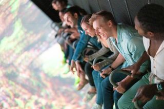 Men and women smile during a Flyover ride in front of a giant screen