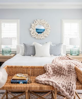 A blue bedroom with a blue wall with white mirror, white bed, rattan bench, and knitted throw