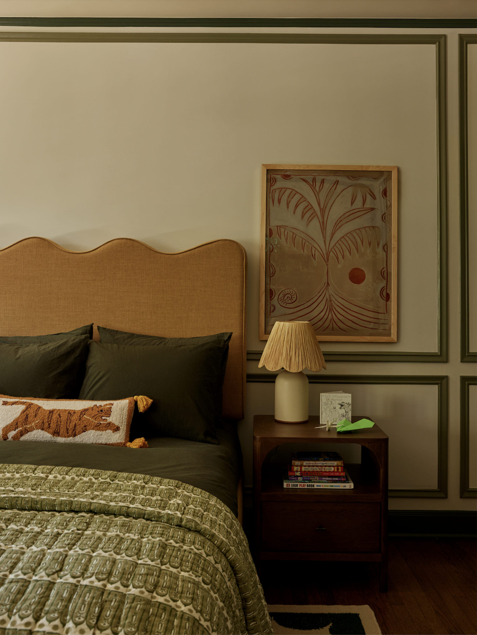 A bedroom with wainscoting, a wavy ochre headboard, green bedding, and a styled nightstand