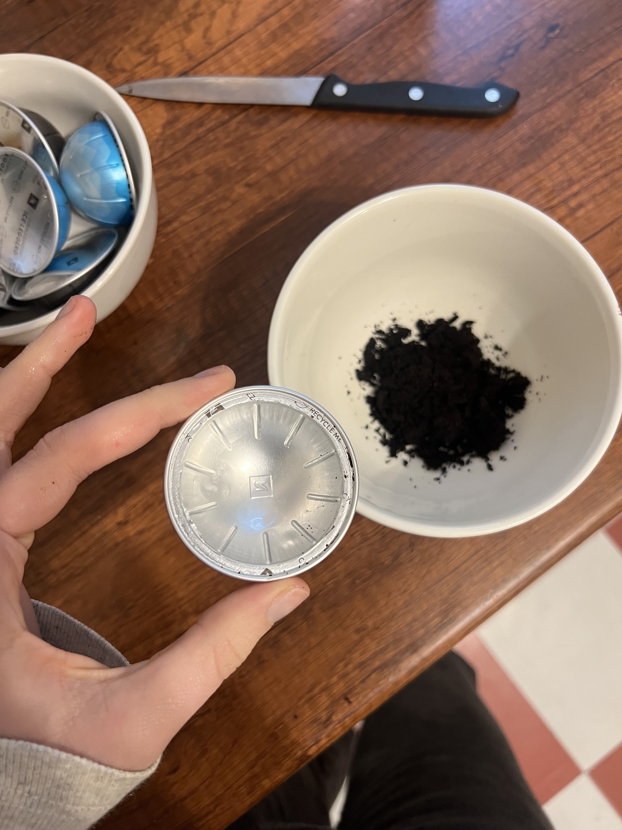 image of a hand holding an open coffee pod that's been emptied over a wooden countertop where there is a bowl full of the discarded grounds. There is a bowl of coffee pods also on the counter.